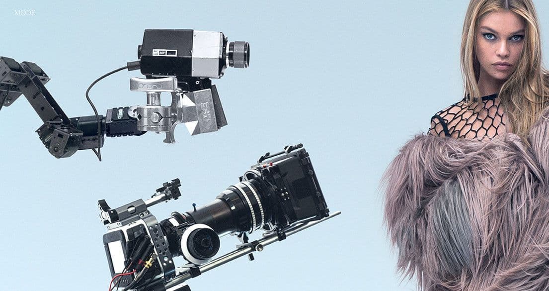 Blonde model in a fishnet top and fur coat standing beside robotic camera arms and film cameras