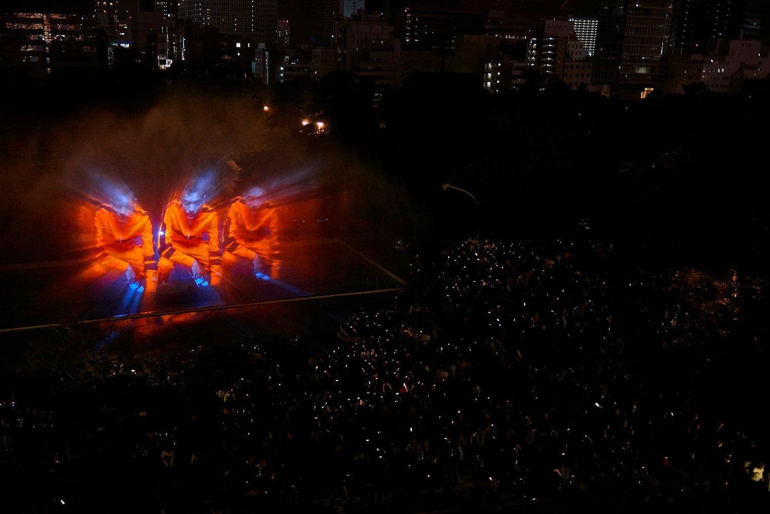 Aerial view of an illuminated outdoor stage with orange and blue lighting, surrounded by a large nighttime crowd.
