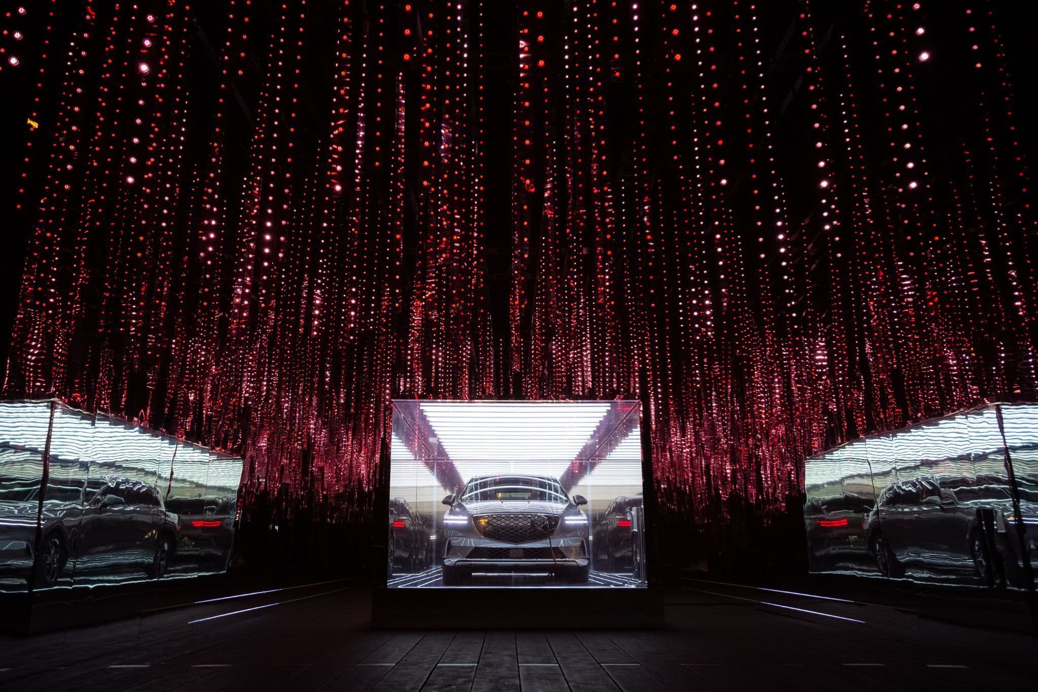 A car centered in a boxy mirrored frame against a backdrop of hanging red string lights.