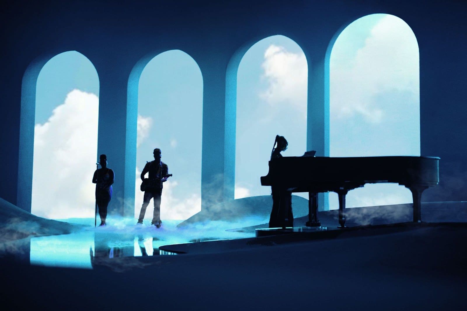 Silhouetted musicians walking between glowing blue arched stage doorways with cloud projections.