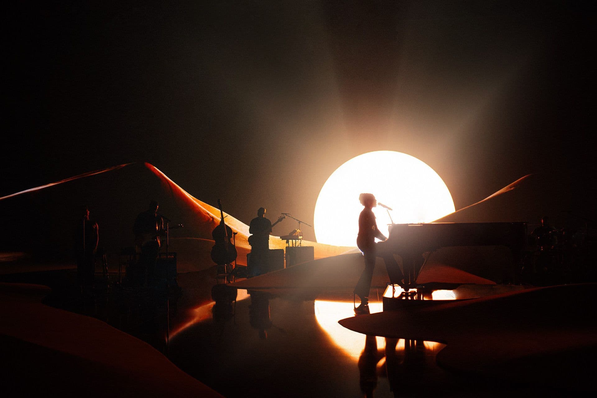 Silhouetted performer at a piano on a dune-shaped stage with a large glowing sun behind, band members in shadow.