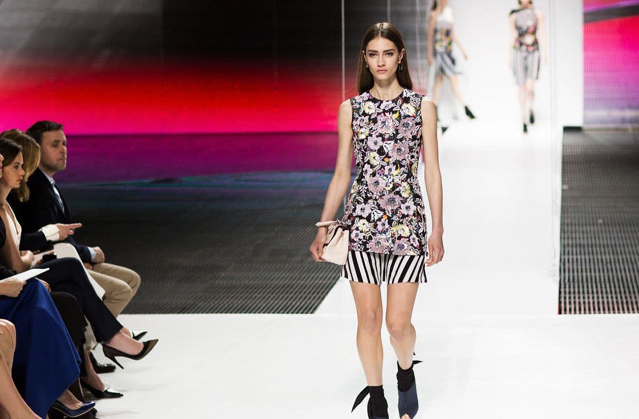 A model in a black-and-white floral print top and striped skirt walks past seated front-row guests, with a pink-lit backdrop.