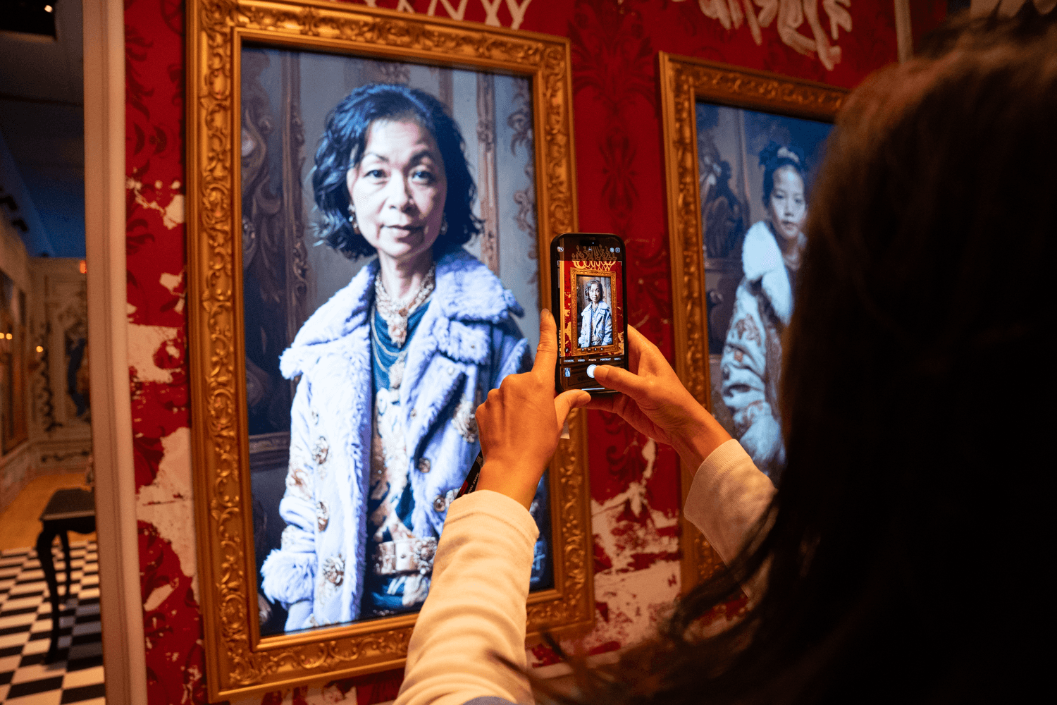 A visitor photographs a framed portrait on a red gallery wall using their phone.