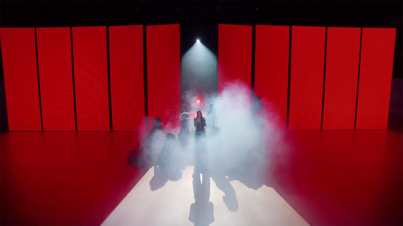 Silhouetted figures walk through fog on a runway lined with tall red panels and red lighting.