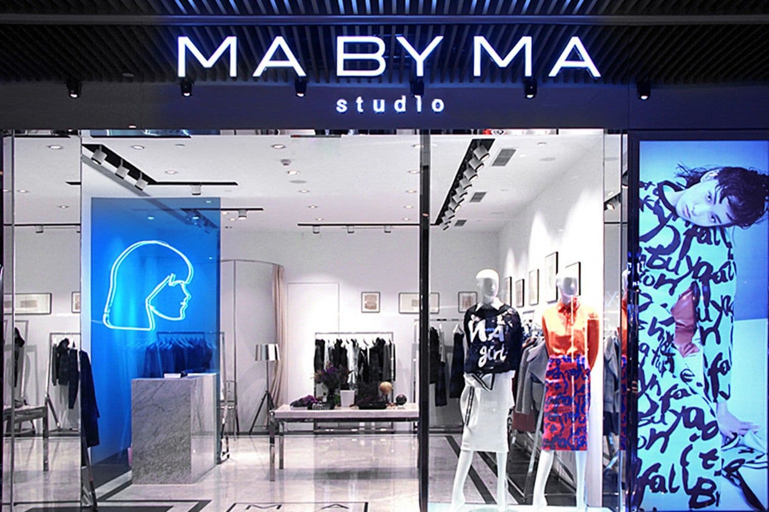 MA BY MA studio storefront at night with an illuminated sign, a blue neon profile logo, and mannequins in patterned outfits in the window.