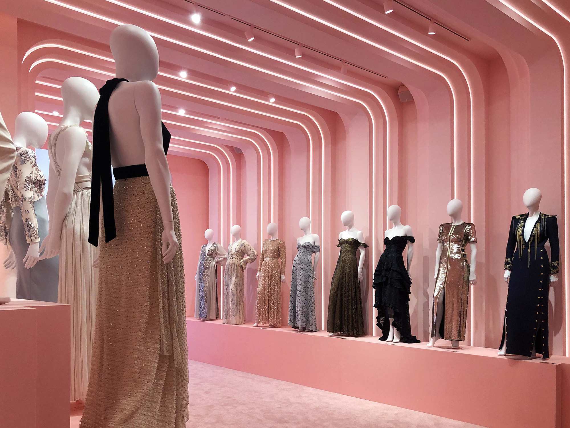 Curved pink corridor with a mannequin in a sleeveless gown in the foreground and dressed mannequins lining the wall.