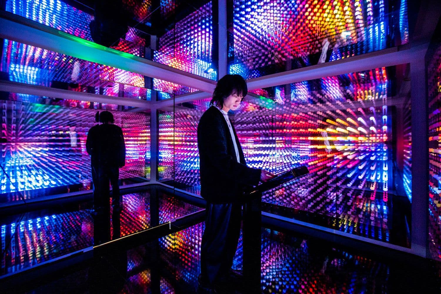 Silhouette of a person checking a phone inside a mirrored room lined with multicolored LED panels.