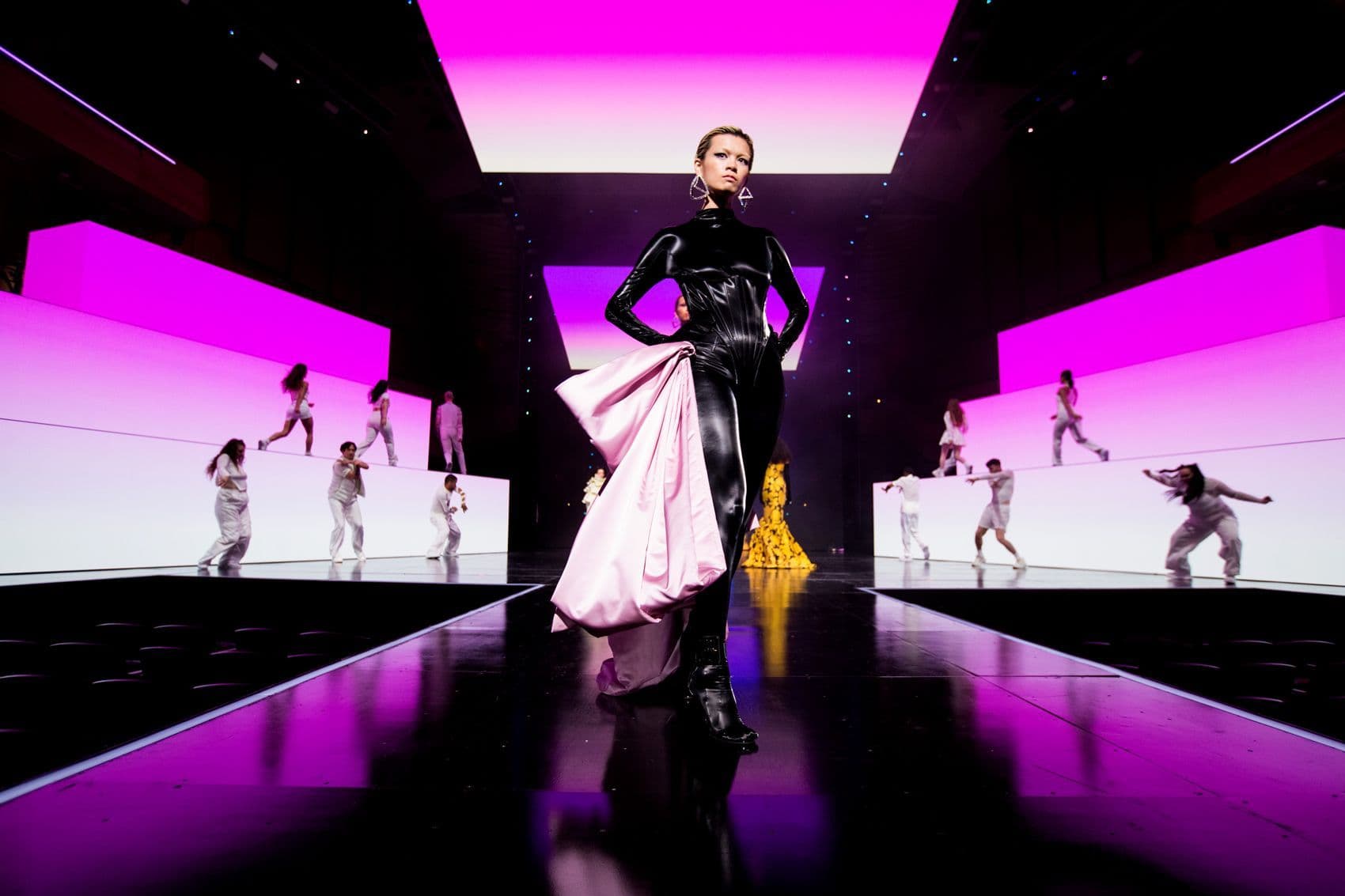 Performer in a flowing white and black gown walking a runway-style stage with dancers, lit in magenta