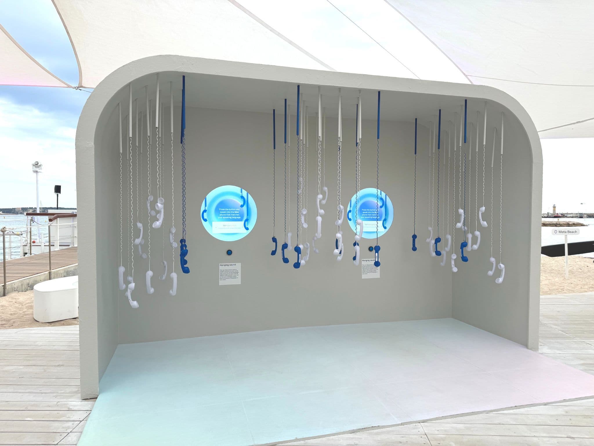 An arched alcove with rows of corded telephone receivers hanging from the ceiling around two illuminated circular blue screens, set on a beach boardwalk.