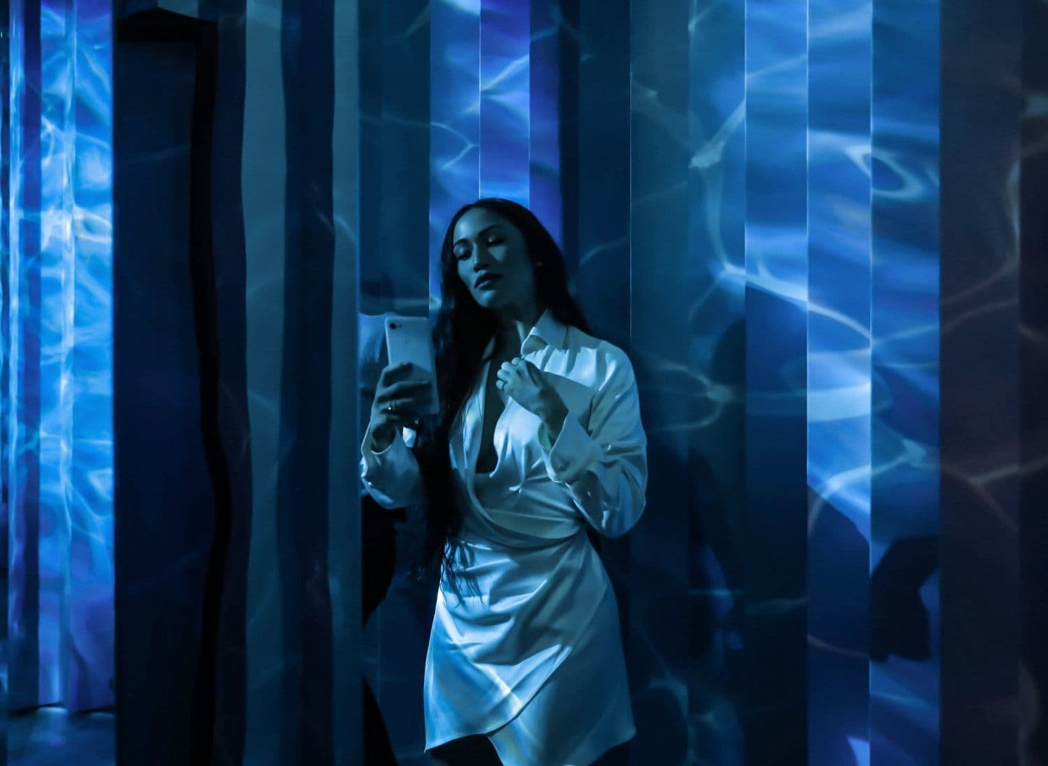 A woman in a silver jacket photographing herself against a blue-striped light installation.