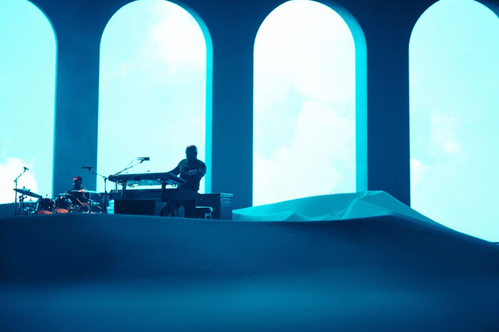 Silhouetted musician at a keyboard beneath glowing blue arched stage set pieces.