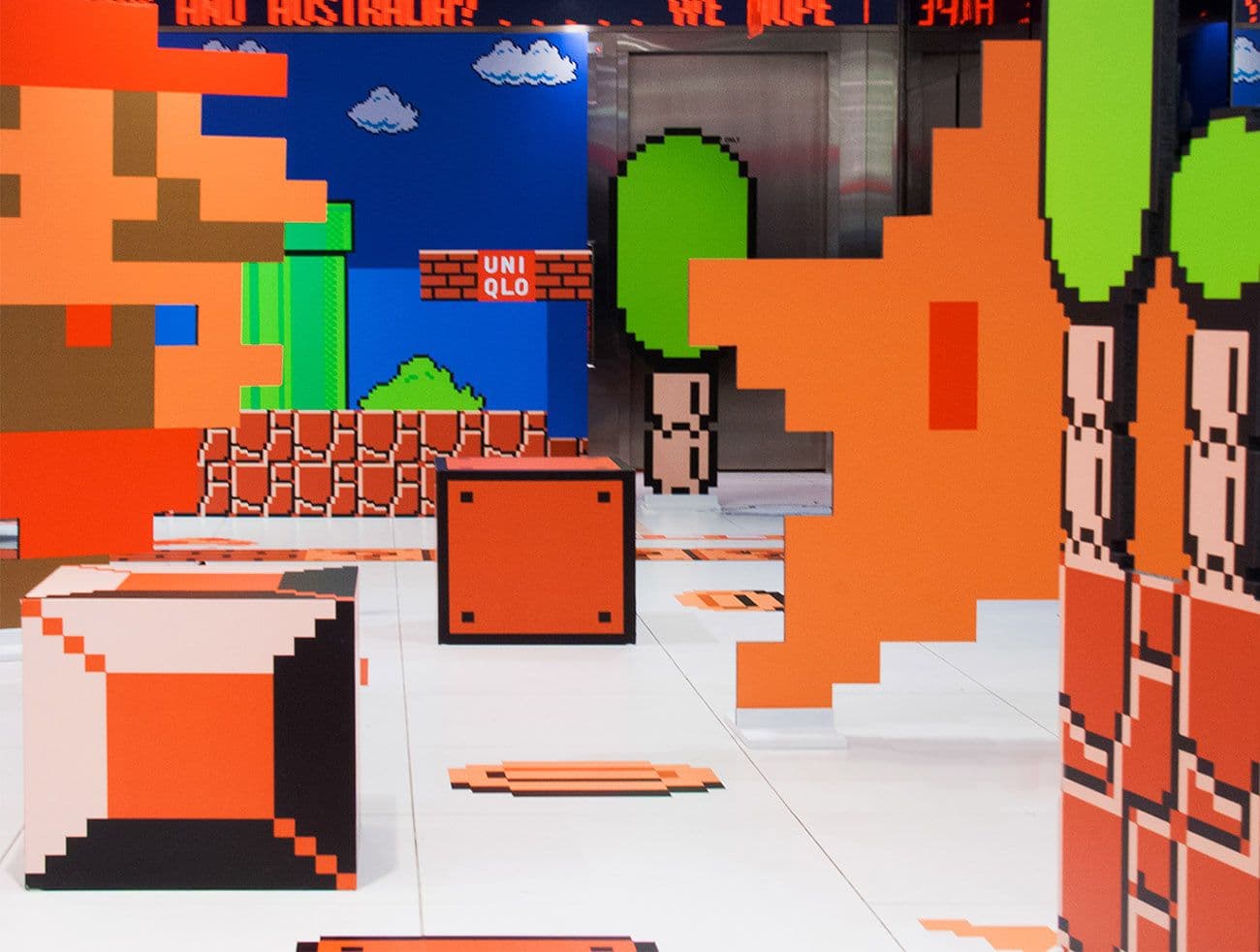 White gallery space with oversized orange pixel-art block sculptures and a Super Mario-themed wall mural, UNIQLO logo on the floor.