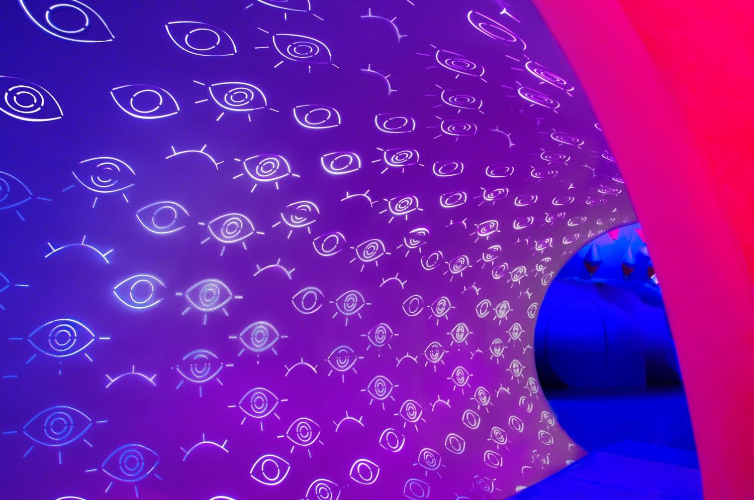 Curved purple ceiling and wall densely covered in illustrated eye patterns, backlit purple and blue.