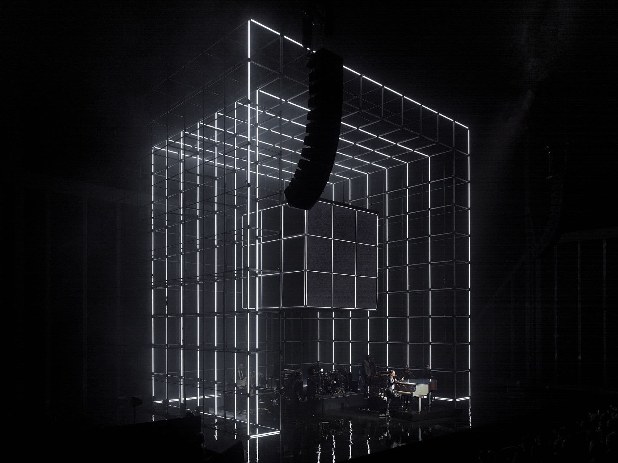 Illuminated wireframe cube-shaped stage structure with a dark video panel at its center, set up over a piano and band area in an empty dark venue.