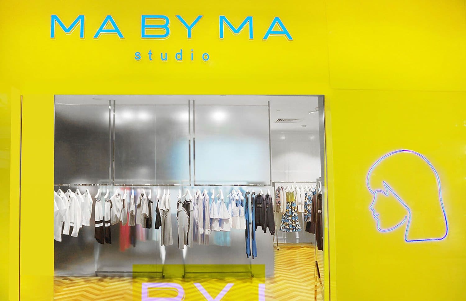 Yellow MA BY MA studio storefront with a neon profile logo beside the entrance and clothing racks visible through the glass.