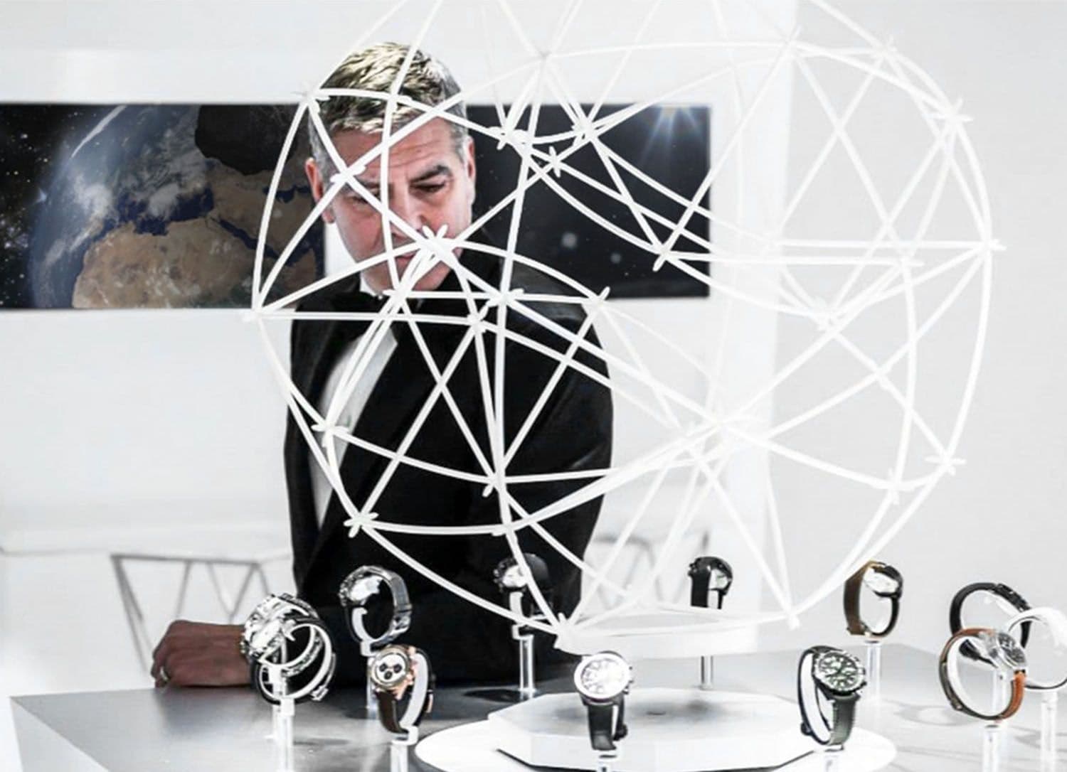 Man in a tuxedo examining a table of watches inside a spherical wireframe sculpture, with an image of Earth on the screen behind him.