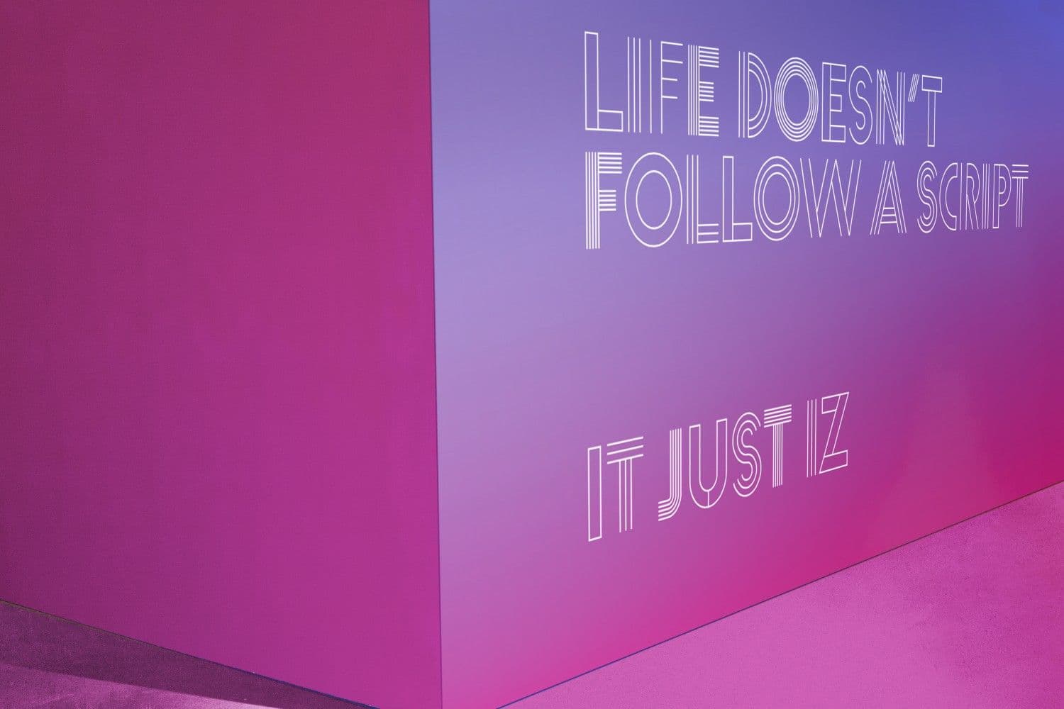 Close-up of a pink wall painted with the text "LIFE DOESN'T FOLLOW A SCRIPT / IT JUST IZ".