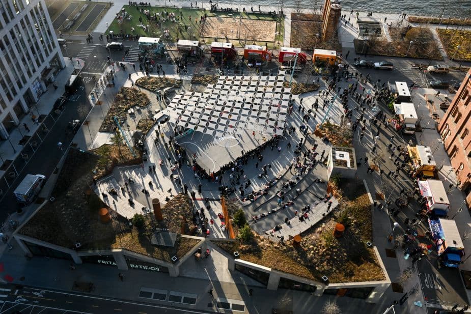 Aerial view of a geometric tented pavilion structure set up in a parking lot alongside city streets.