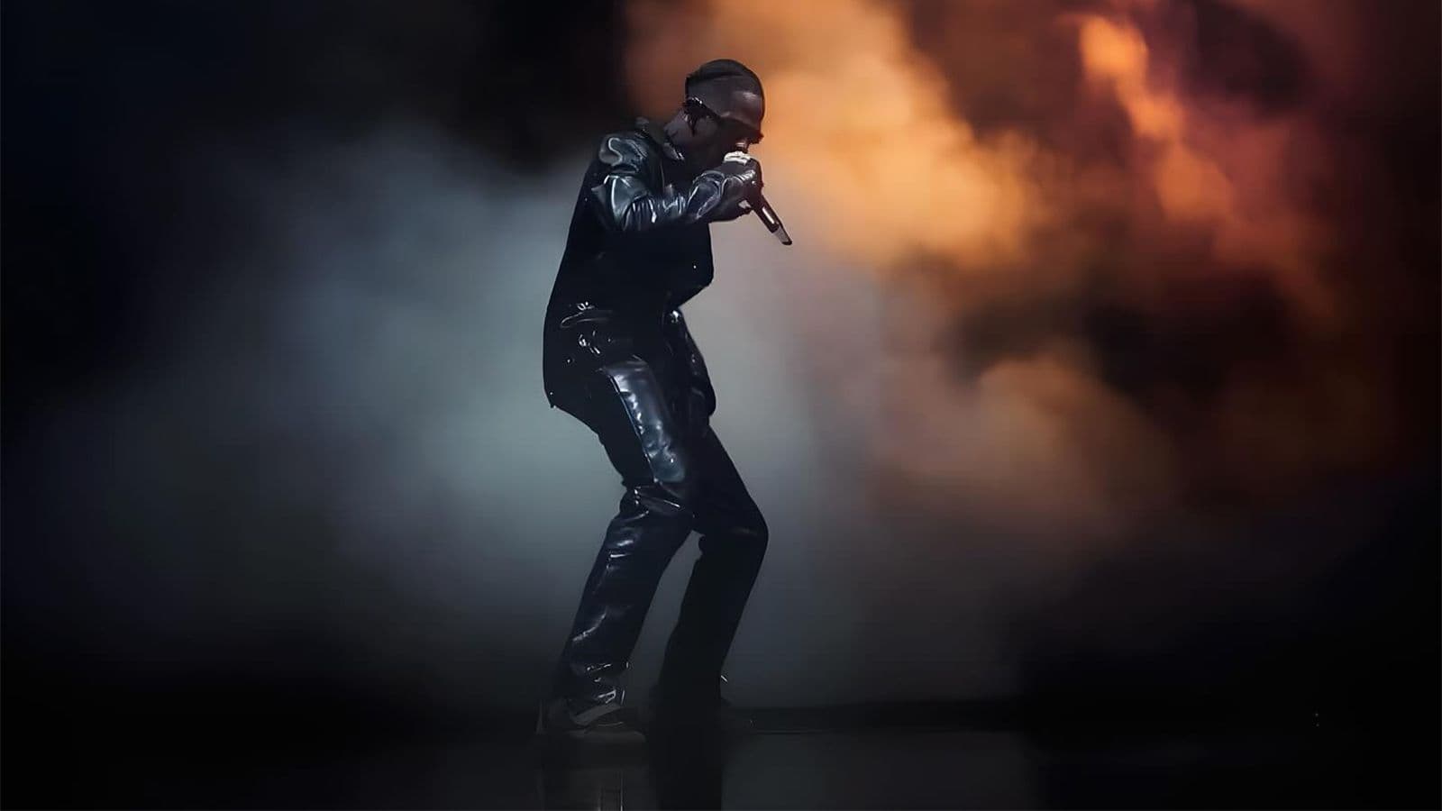 Performer crouched low, singing into a microphone against a backdrop of orange flames.