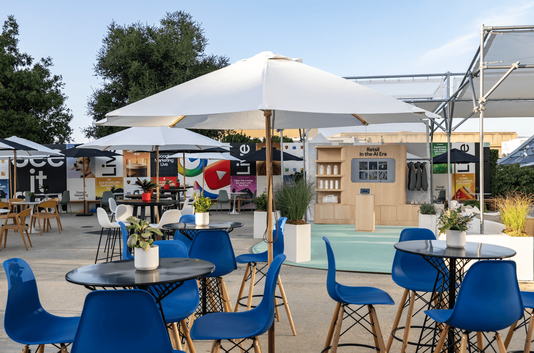 Outdoor lounge area with white umbrellas, round tables, and blue chairs near an event kiosk.