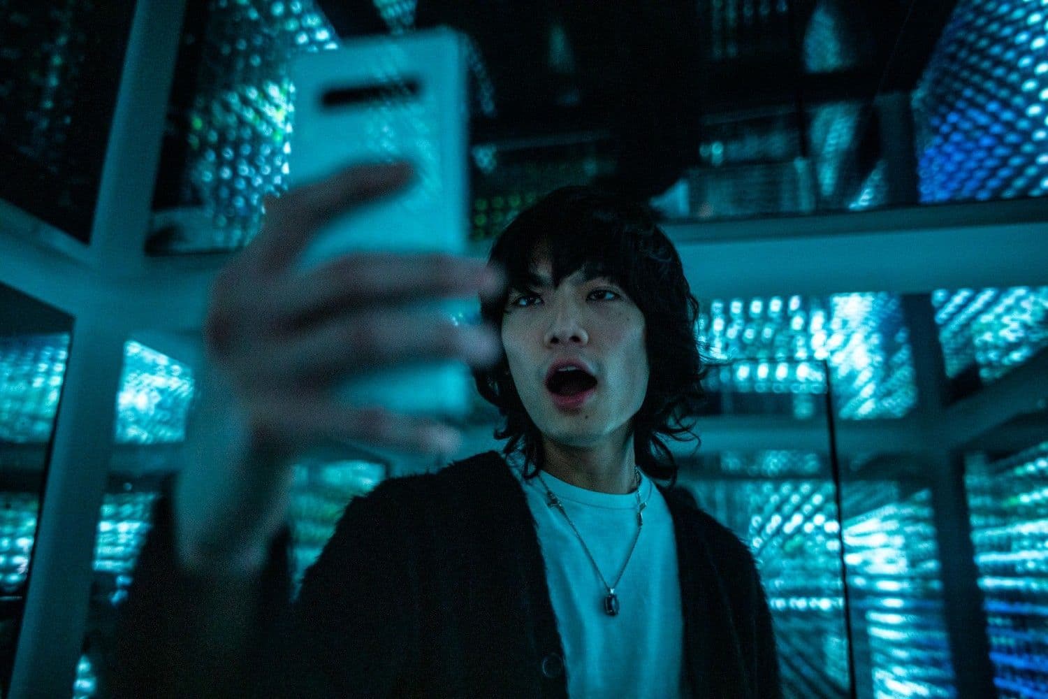 A person reaches upward with mouth open inside a teal-lit LED tunnel of mirrored panels.
