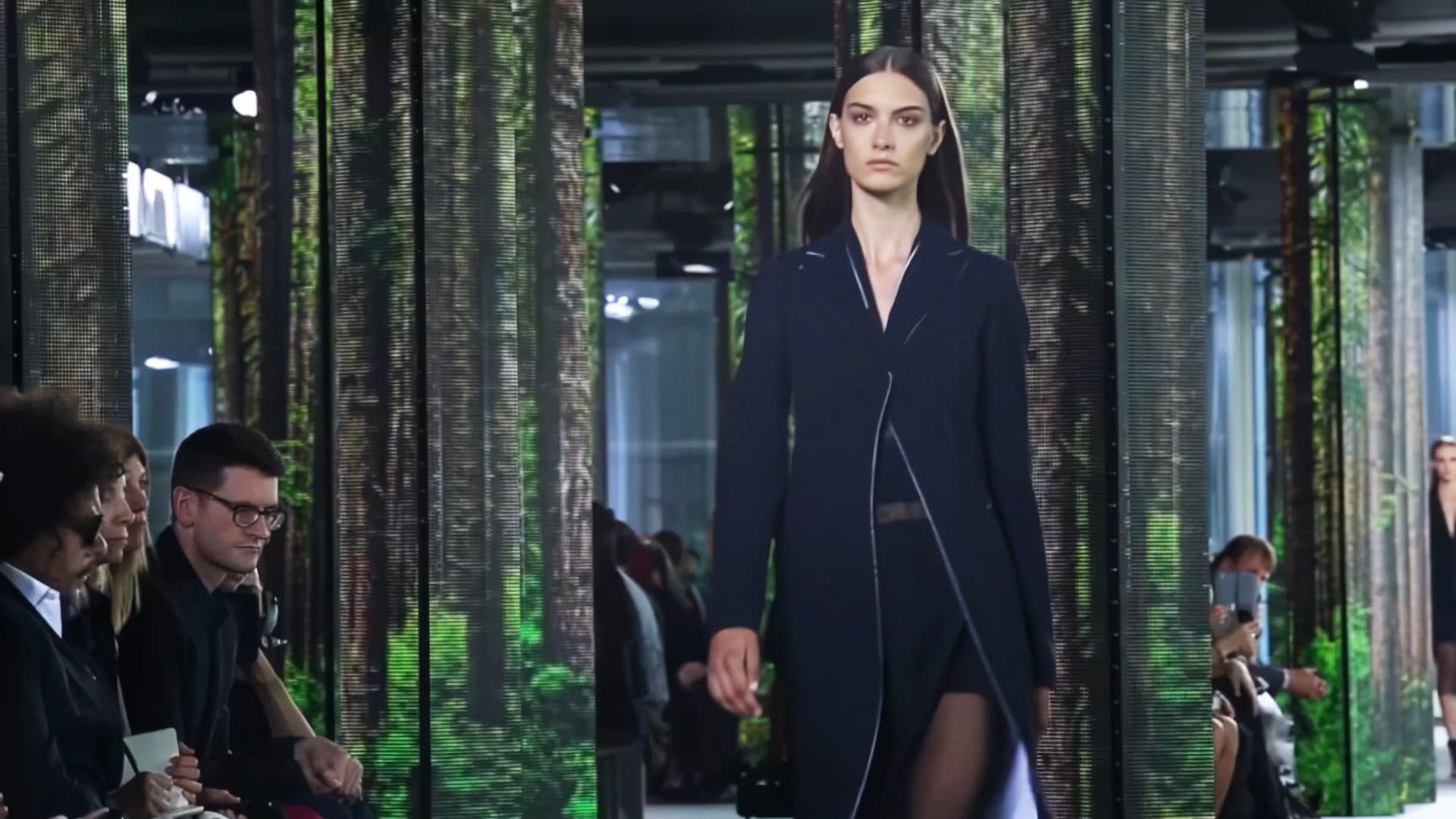 Model in a navy coat walking the runway past a seated audience among forest-print LED columns.
