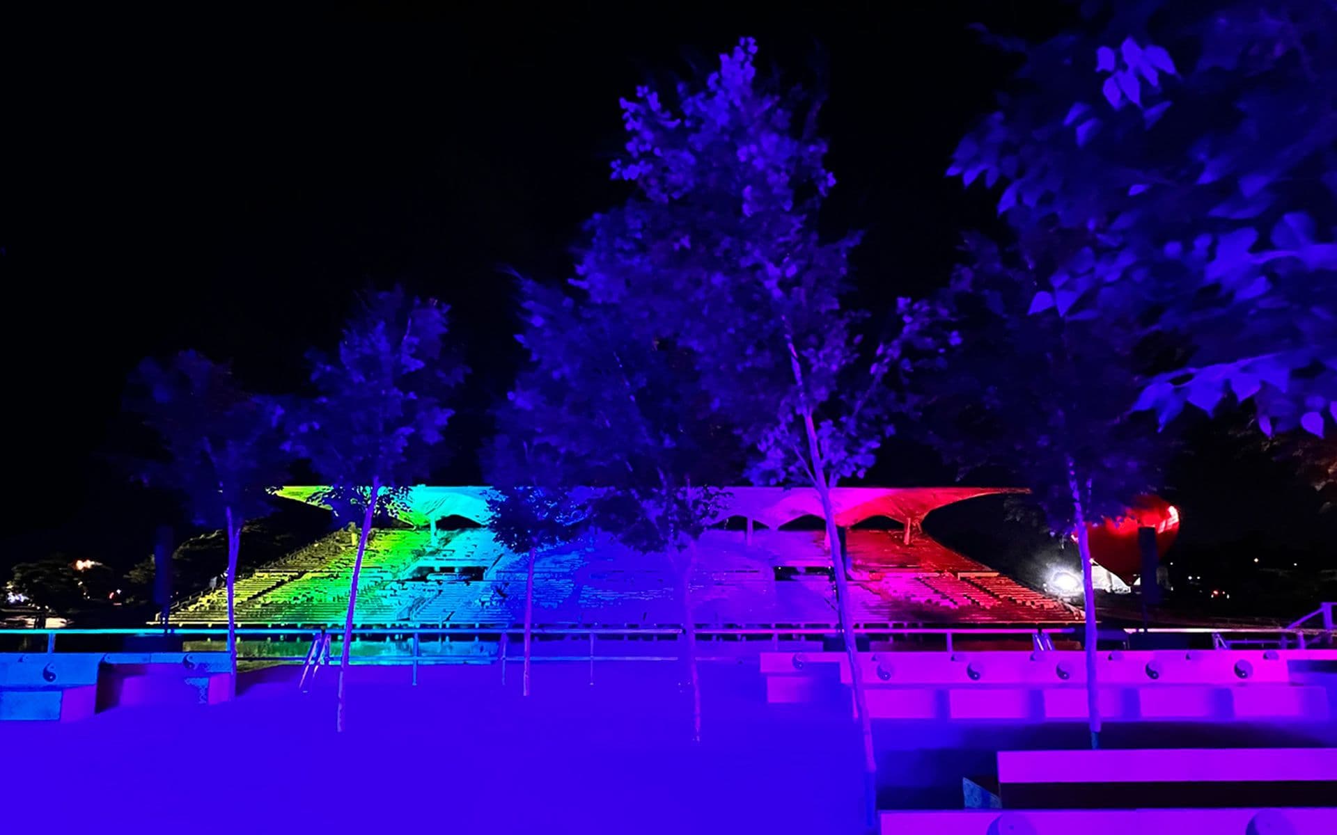 Rainbow-lit installation at night with a red-lit plaza floor and silhouetted trees in the foreground