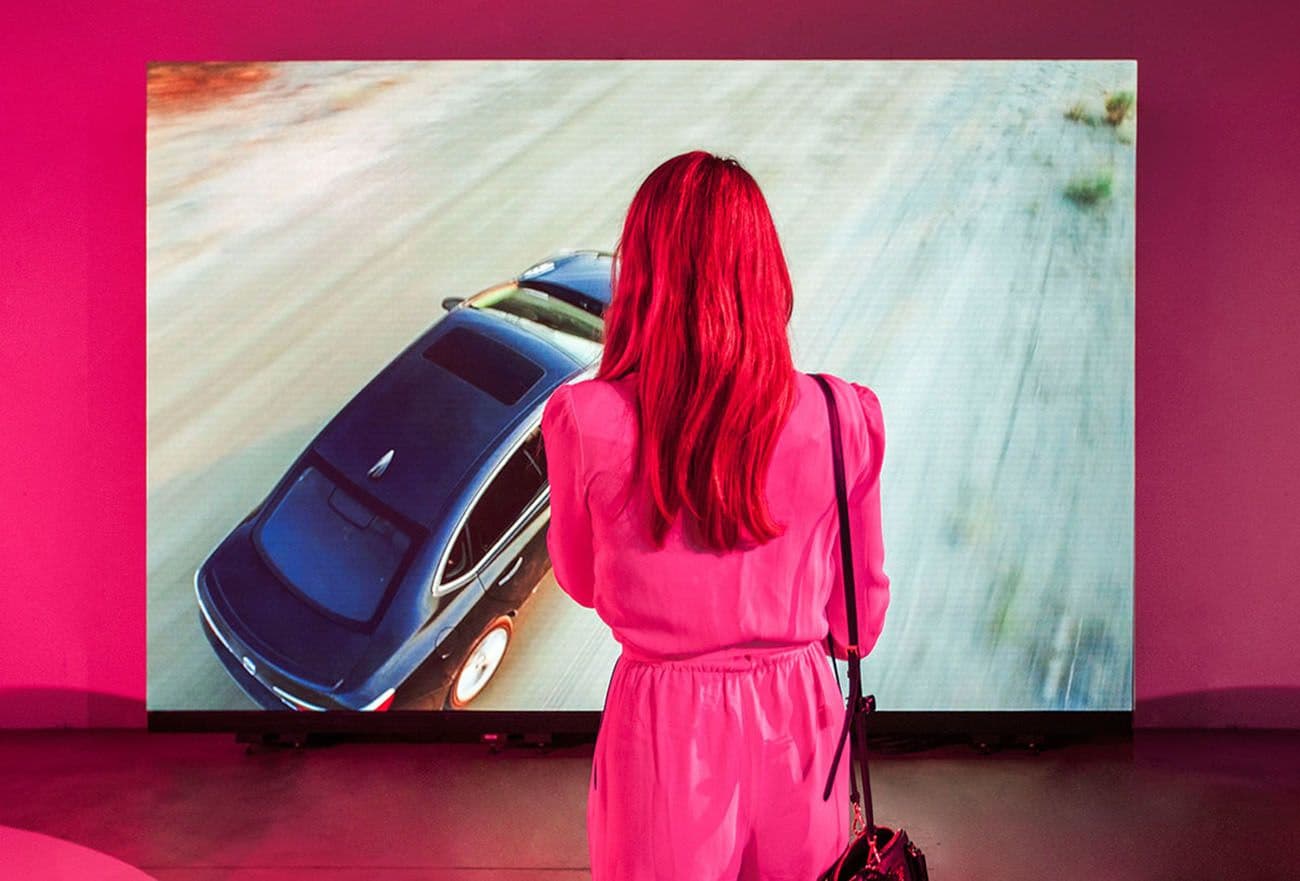 A woman with red hair in a pink dress standing in front of a large screen showing a car driving on a desert road, viewed from behind.
