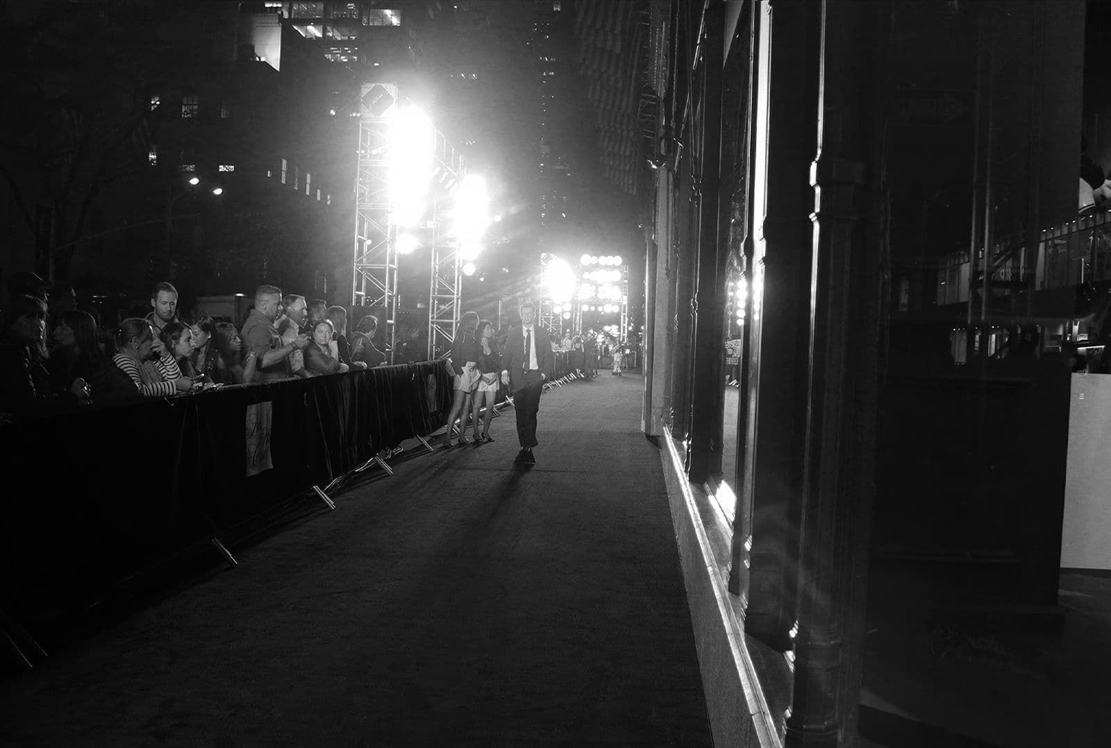 Black-and-white night street scene, a crowd behind a barrier lit by a bright light.
