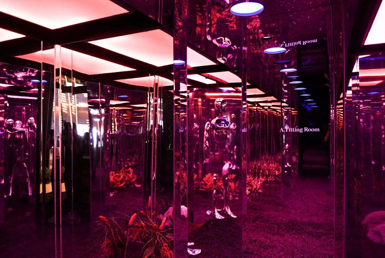 Mirrored room reflecting pink light, greenery, and a sign reading 'Fitting Room' repeated across multiple angled mirror panels.