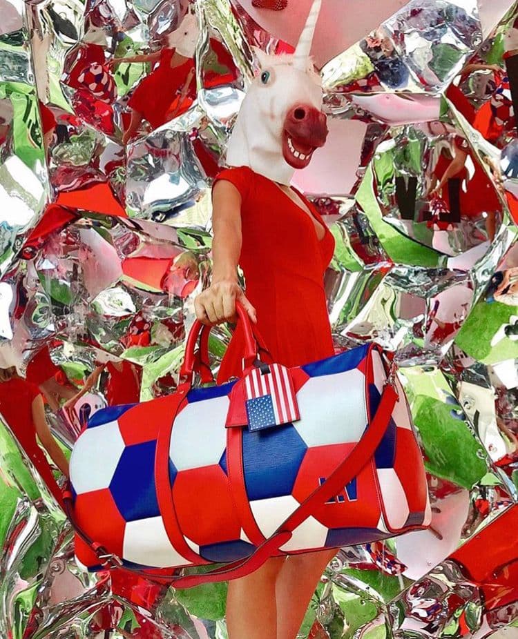 Performer in a unicorn mask and red dress holding a red, white and blue Louis Vuitton Keepall bag against a mirrored mosaic backdrop.