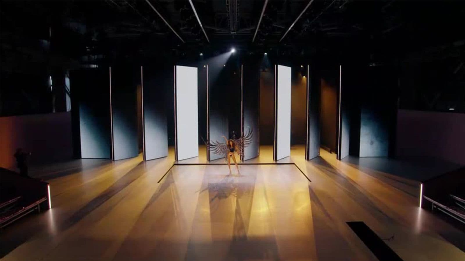 A model walks the runway between two rows of tall backlit panels in a warmly lit venue.
