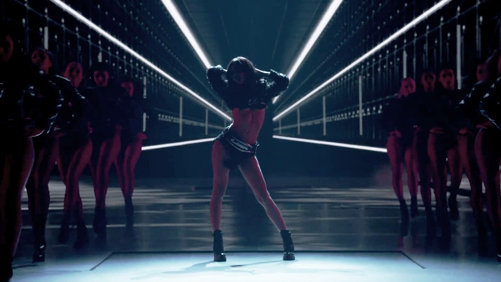 A model in a black bodysuit dances on the runway in a narrow, brightly lit corridor flanked by silhouetted dancers in red masks.