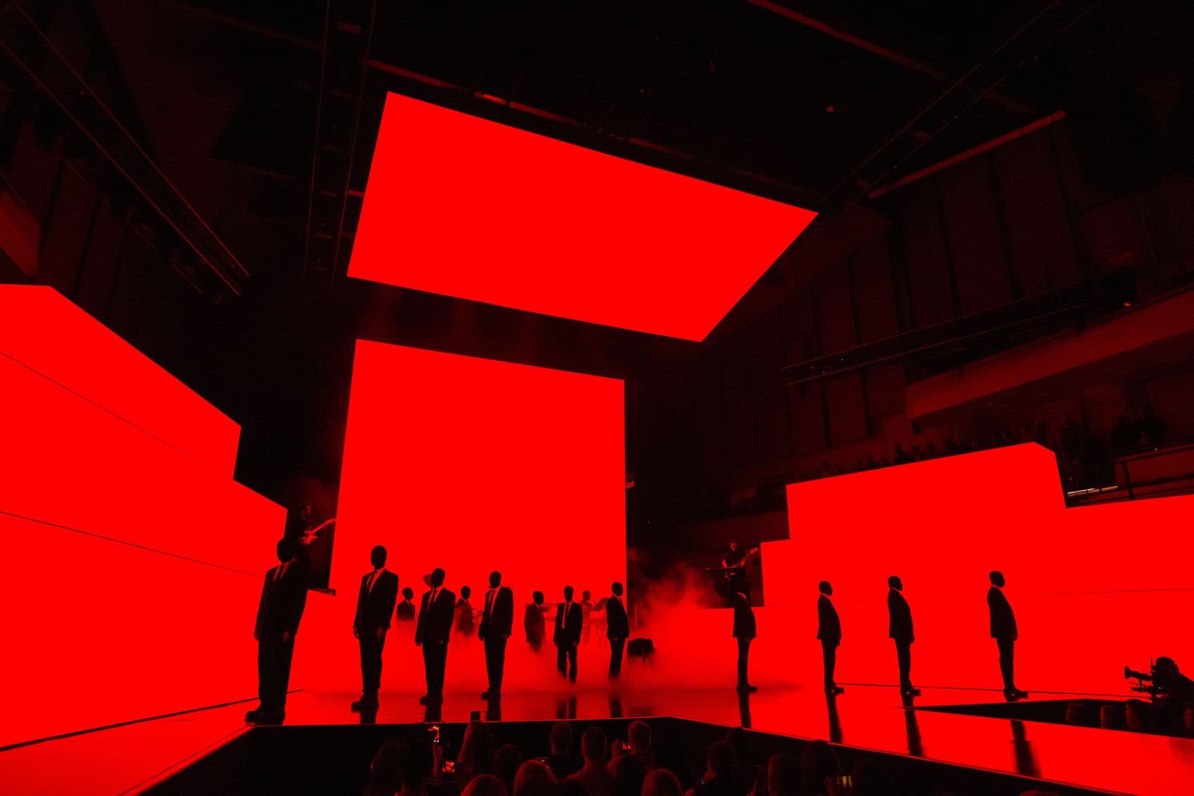 Line of silhouetted people standing against a red backlit angular stage structure.