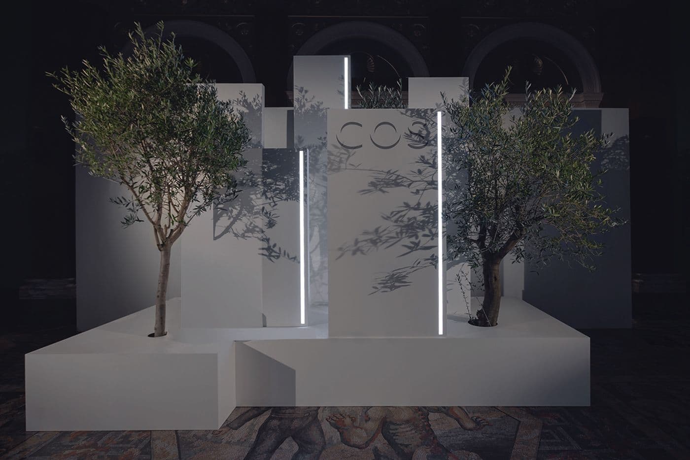 White pedestal installation with two potted olive trees and illuminated COS lettering, staged on a mosaic floor inside a darkened museum hall.
