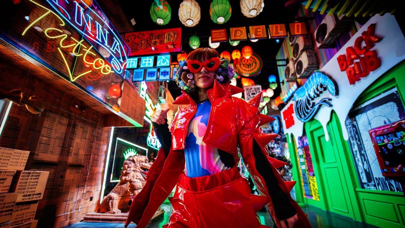 A figure in a red devil-horned costume poses in a neon-lit alley set with signage reading Sushi and other illuminated shop signs.
