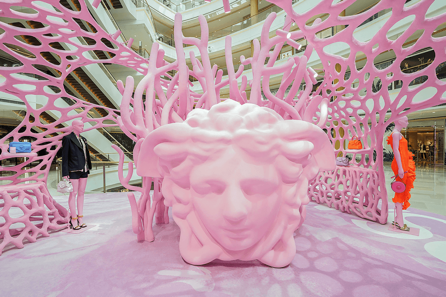 Wide view of the pink coral-branch pavilion inside a shopping mall atrium, with a pink Medusa head sculpture at its center and mannequins in black and orange outfits nearby.