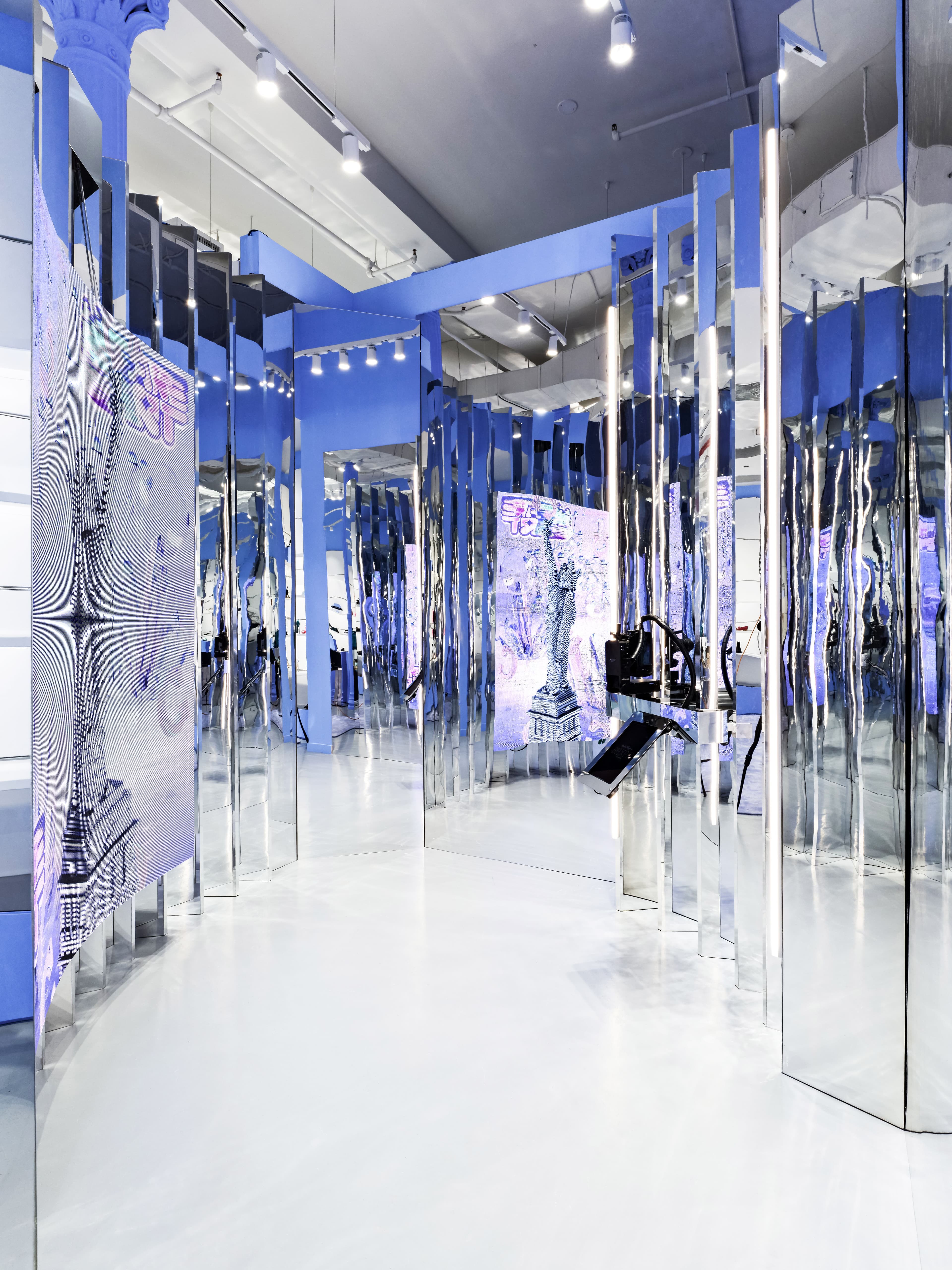 Mirrored store corridor with cobalt-blue graphic wall panels of an illustrated sculpture, a shopper reflected walking through.