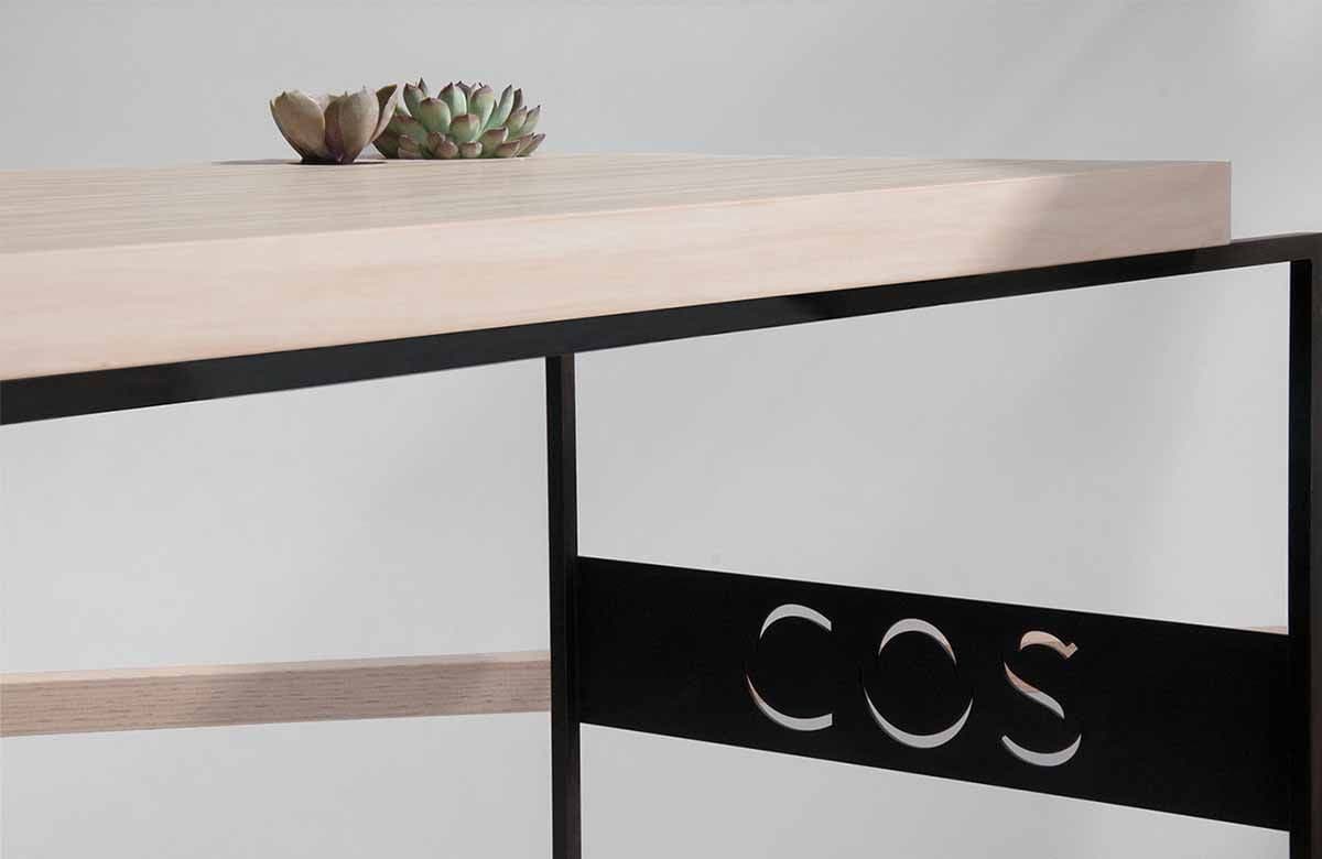 Close-up of a wood tabletop edge with a succulent above a black metal panel with the COS logo cut out.