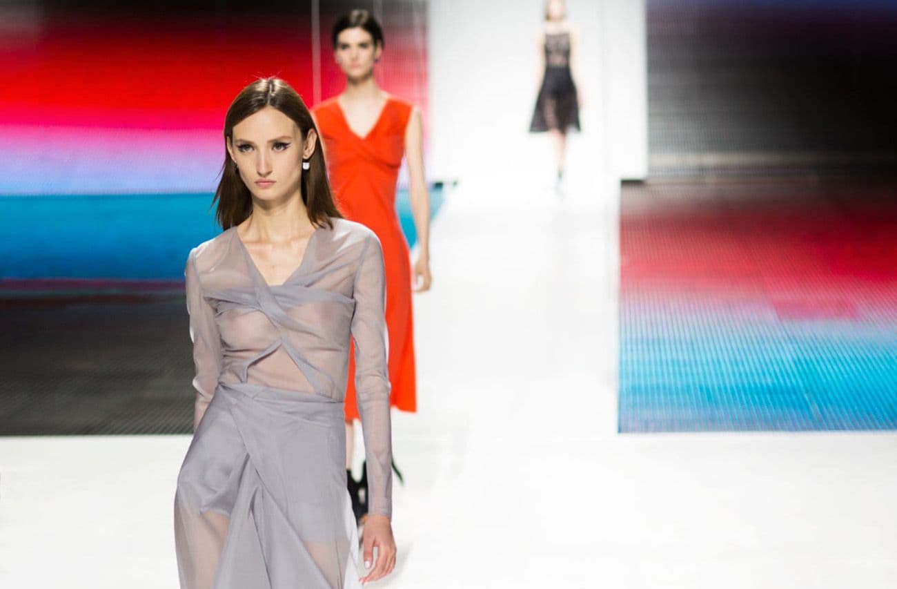 Two models walk the runway, one in a sheer grey wrap dress and the other in a burnt-orange dress, against a blue gradient backdrop.