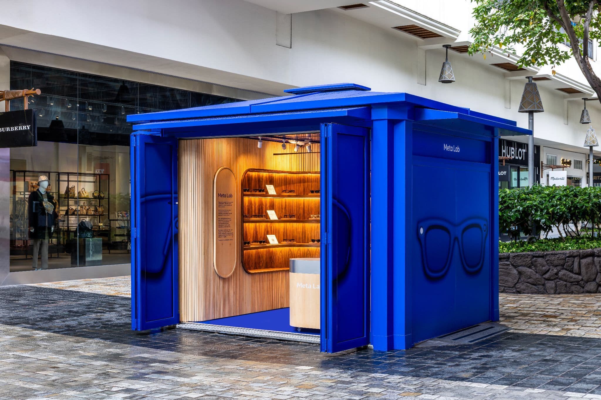 Small blue freestanding kiosk with its front panel opened outward, revealing wood shelving inside, set on an outdoor plaza.