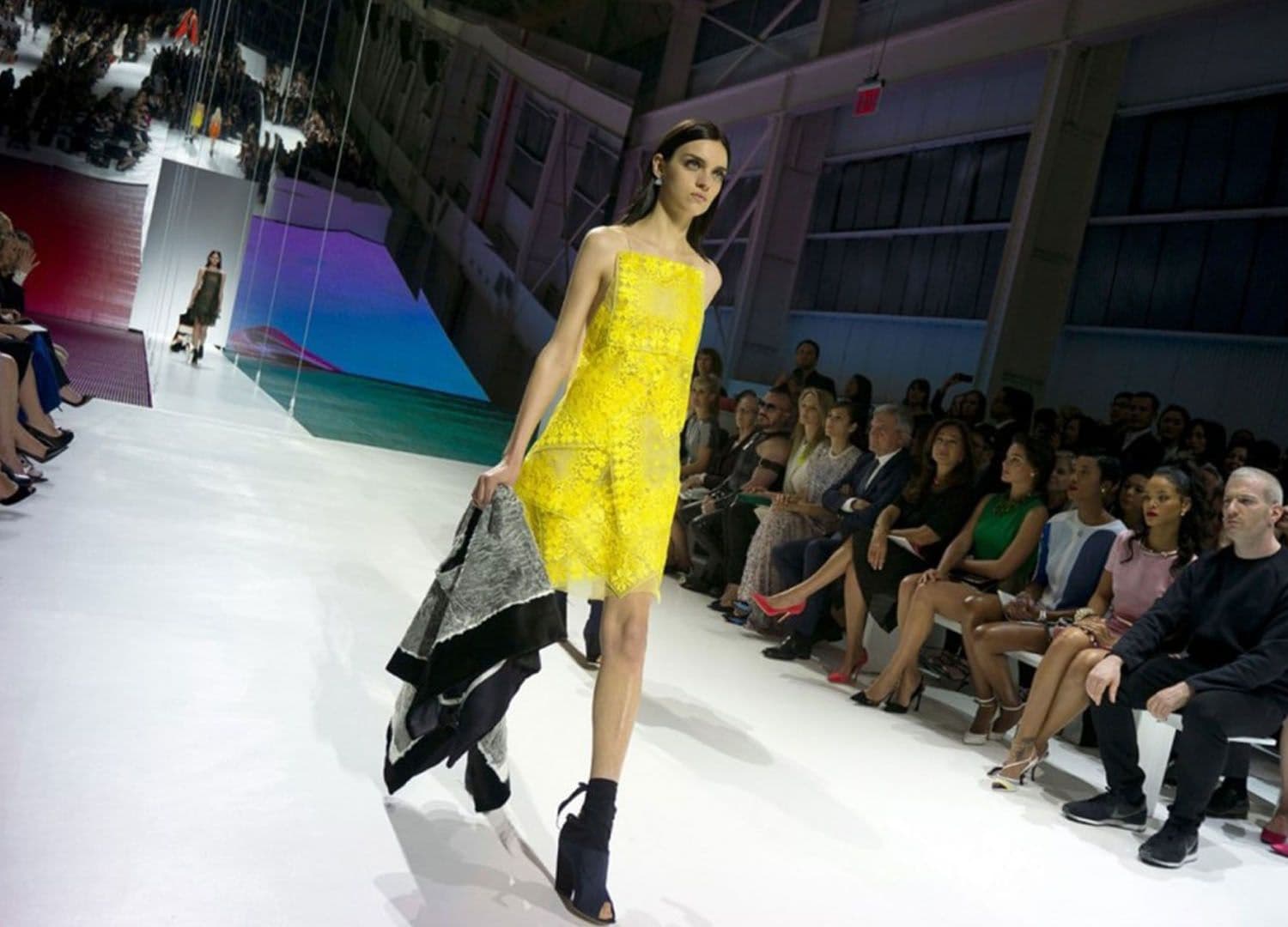 A model in a mustard-yellow coat and black skirt walks past seated audience members on a striped runway floor.