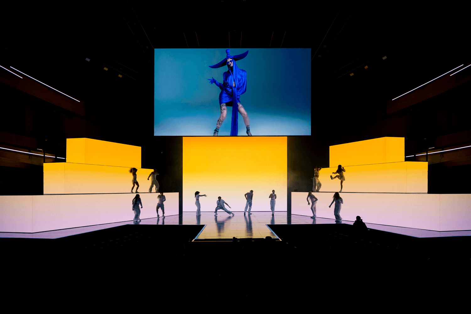 Dancers performing on illuminated yellow platforms with a screen showing a performer above