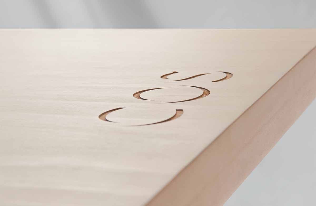 Close-up of the COS logo engraved into a pale wood tabletop corner.
