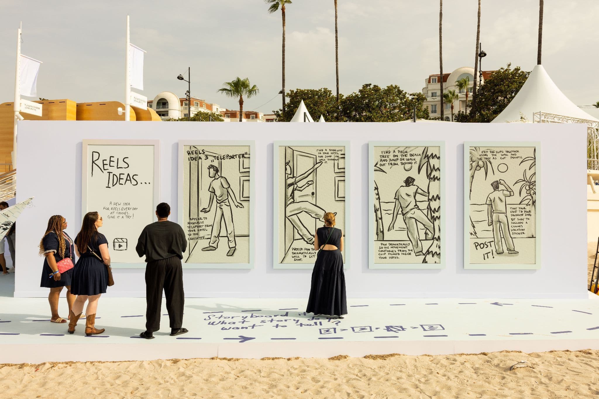 Visitors look at a row of illustrated comic-style panels titled 'Reels Ideas' mounted on a white wall near a beach, with palm trees in the background.