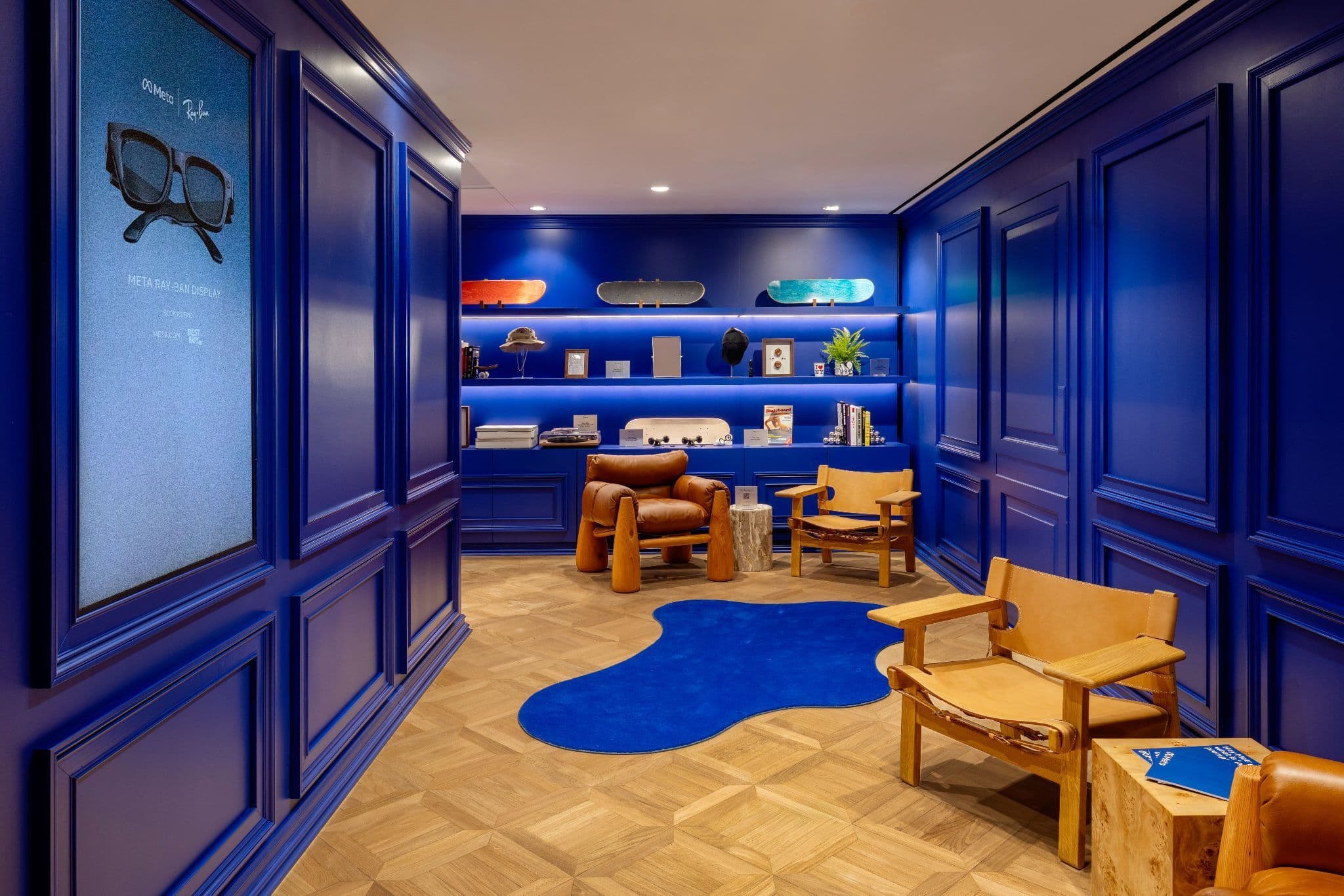 Navy-paneled room with a wall-mounted sunglasses display, wood shelving, a small table and chairs, and a blue amoeba-shaped rug.