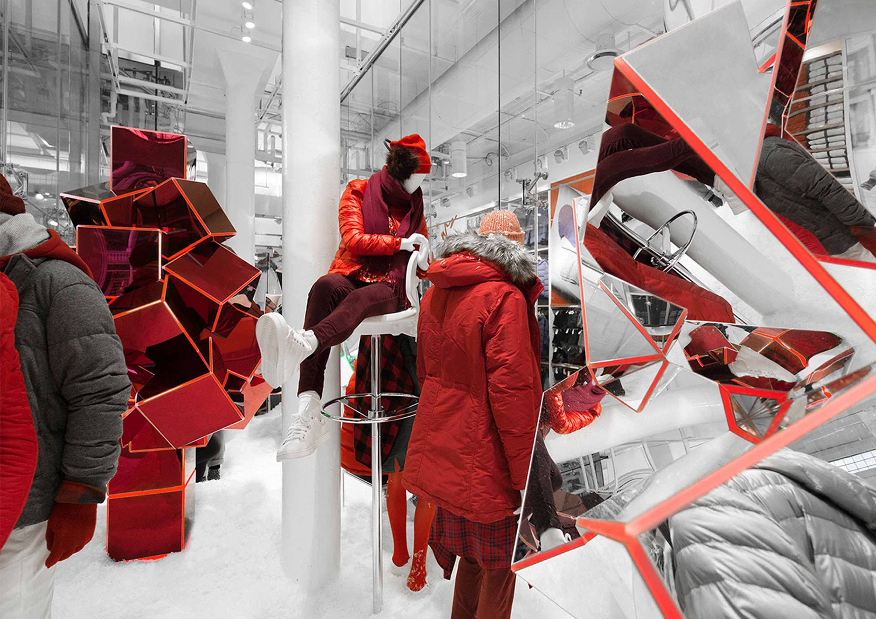 A person in a red mask and coat sits on a stool beside a faceted red-and-mirror sculpture, next to mannequins dressed in red winter coats in a snow-themed store display.